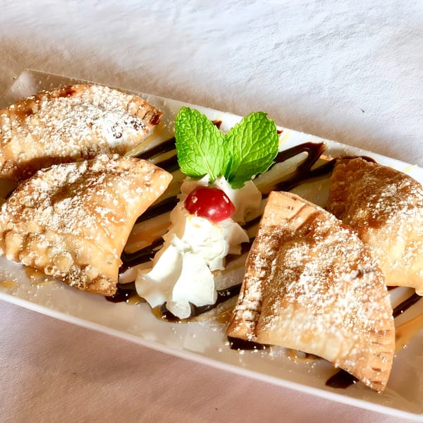 Four golden-brown Cuban dessert empanadas dusted with powdered sugar, garnished with whipped cream, a cherry, and mint.