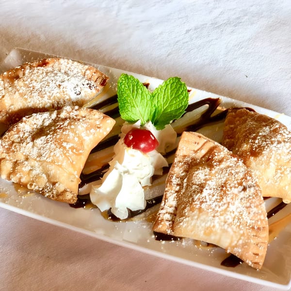 Four golden-brown dessert empanadas dusted with powdered sugar, drizzled with chocolate, and garnished with whipped cream, a cherry, and mint.