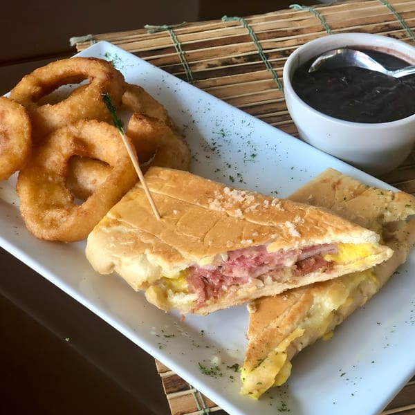 Cuban sandwich with ham, cheese, and pickles, served with onion rings and black beans.
