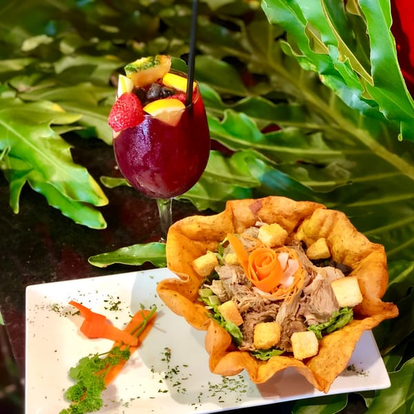 Cuban shredded pork salad in a tortilla bowl with a fruit-filled sangria against lush greenery.