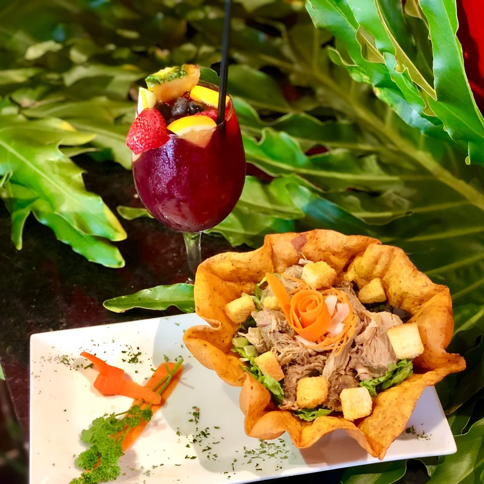 Cuban shredded pork salad in a tortilla bowl with a fruit-filled sangria against lush greenery.