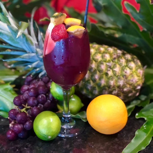 A glass of red sangria garnished with fruit, surrounded by fresh grapes, limes, an orange, and pineapples.