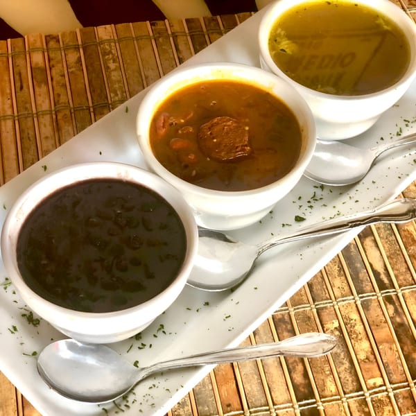 Three small white bowls of Cuban soups: black bean, chorizo, and chicken, on a white tray.