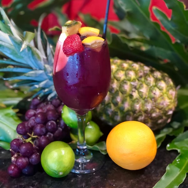 A vibrant purple drink garnished with fruit, surrounded by pineapples, grapes, limes, and an orange.