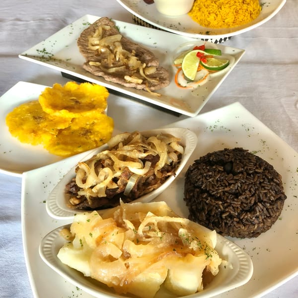 Cuban dishes including steak with onions, plantains, black rice, and yucca on white plates.