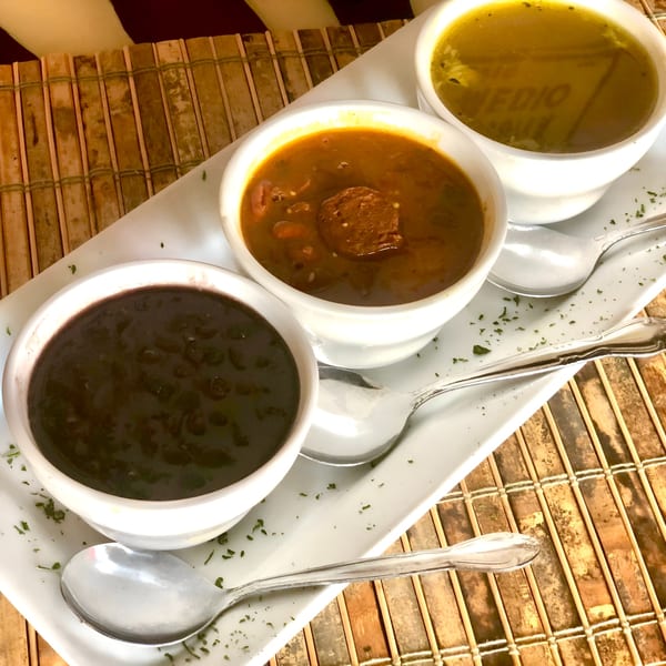 Three small white bowls of Cuban soups, including black bean and a hearty stew, on a white tray.