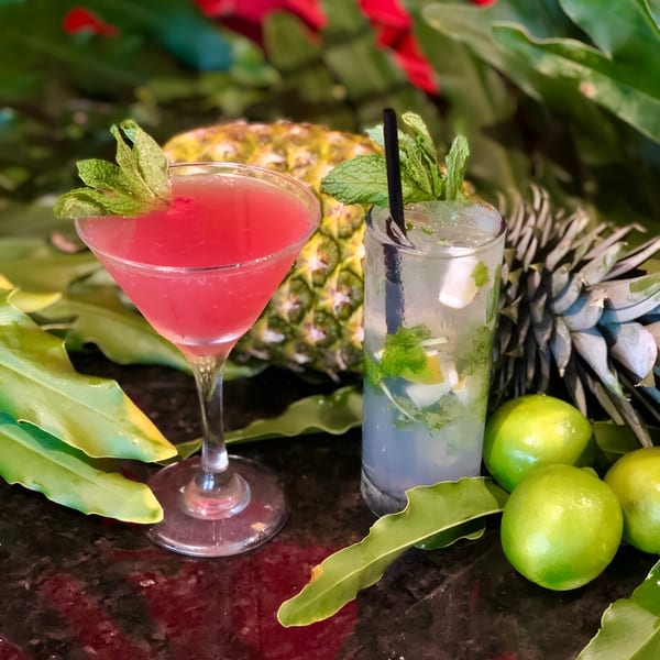 Two Cuban cocktails, a red martini and a mojito, garnished with mint, limes, and pineapple.