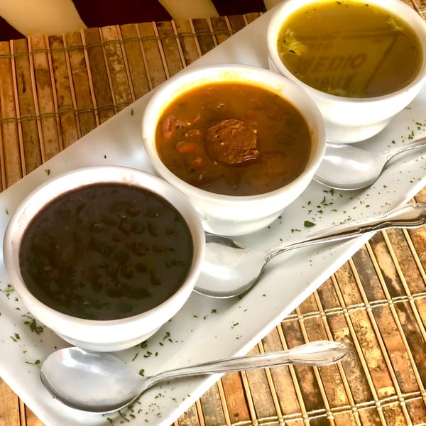 Three small white bowls of Cuban soups, including black bean and a hearty stew, on a white tray.