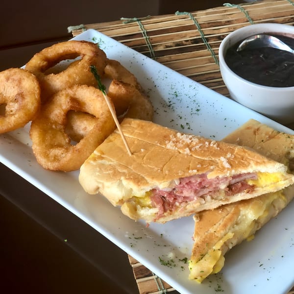 A Cuban sandwich, onion rings, and a bowl of black beans on a white plate at Las Palmas Restaurant.