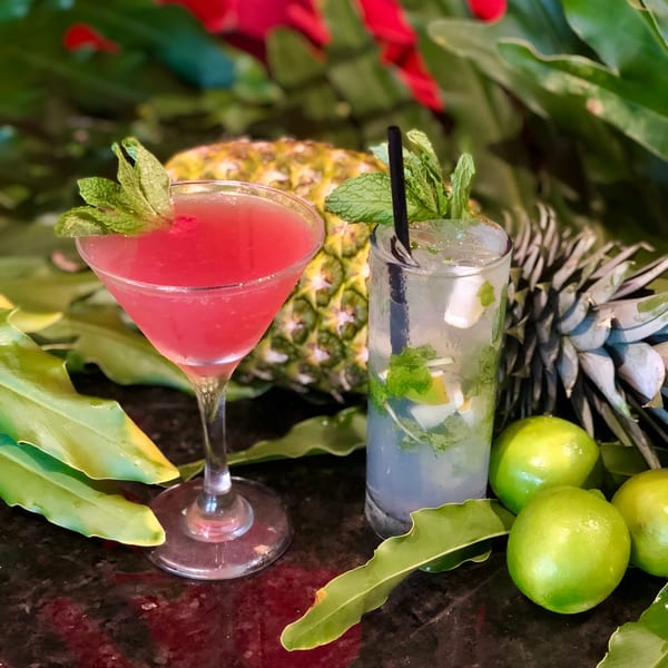 Two Cuban cocktails, a red martini and a mojito, with limes and pineapples on a dark surface.