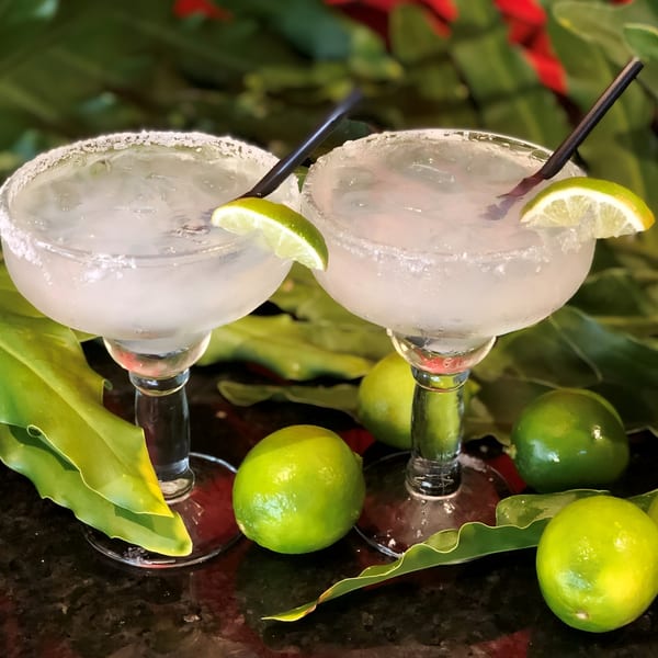 Two frosty margaritas with lime wedges and straws, surrounded by whole limes and green leaves.